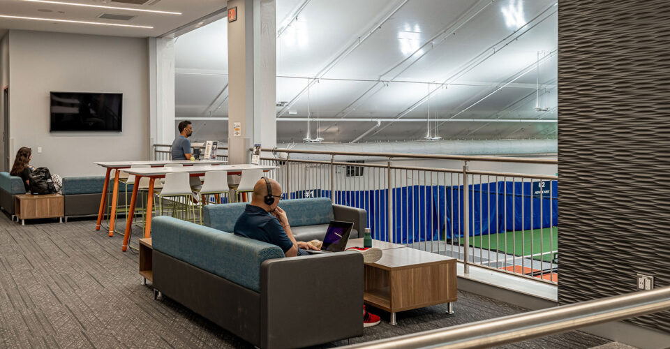 People work on laptops at lounge seating overlooking an indoor sports court in a modern recreational facility.