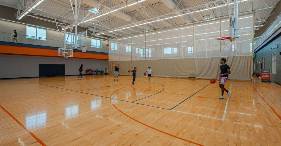 People play basketball on a well-lit indoor court with wooden flooring and high ceilings. Multiple hoops and a few players can be seen spread across the court.