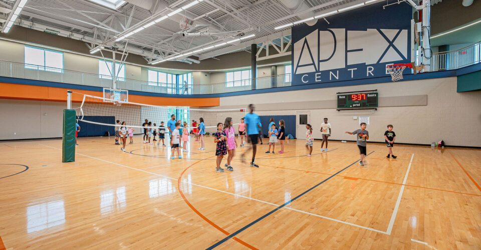 Children and an adult participate in activities on an indoor gymnasium basketball court at the Apex Center, with a volleyball net set up in the background.