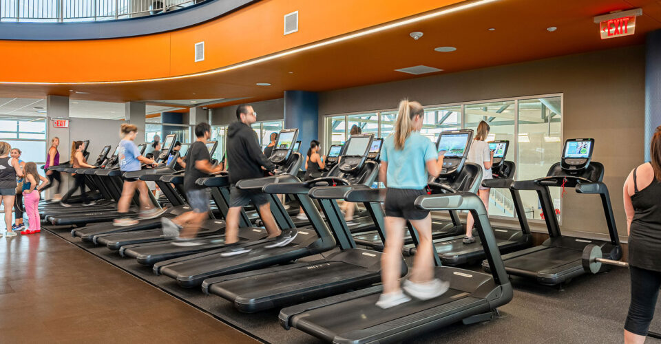 People exercise on treadmills in a modern gym with an orange and gray interior. Some are walking or running while others wait nearby. Large windows provide natural light.