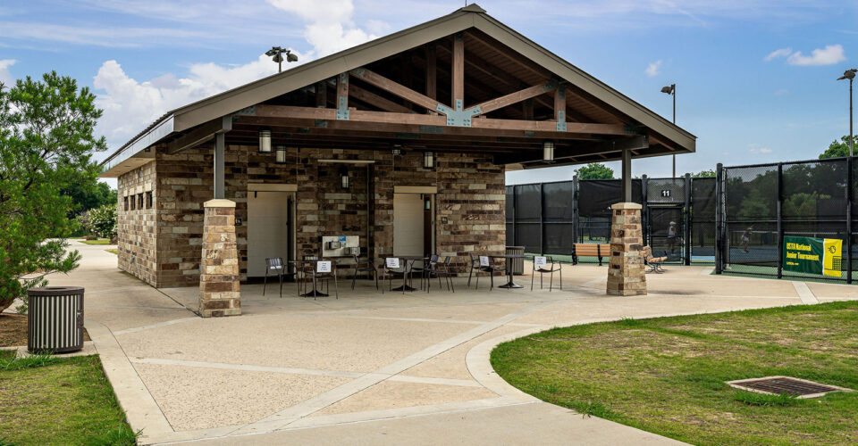 A stone building with a covered patio, tables, and chairs sits beside fenced tennis courts under a partly cloudy sky.