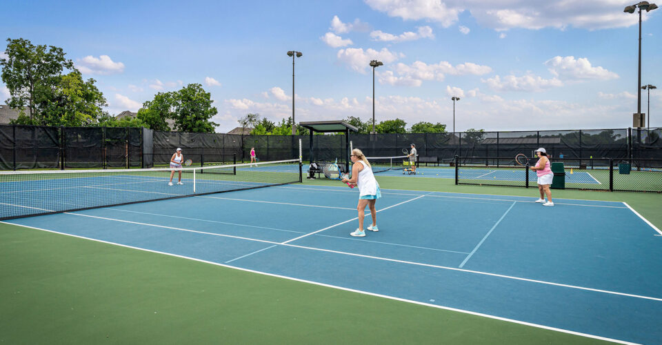 Four people play doubles tennis on an outdoor court under a blue sky with scattered clouds. Trees and black fencing surround the court.