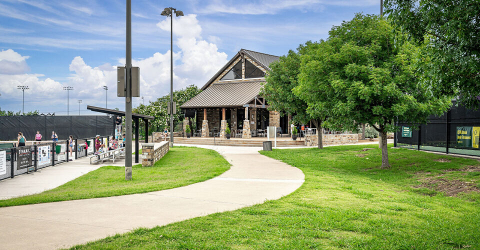 A paved path leads to a stone and wood pavilion surrounded by green grass and trees, with sports fields visible on the left.
