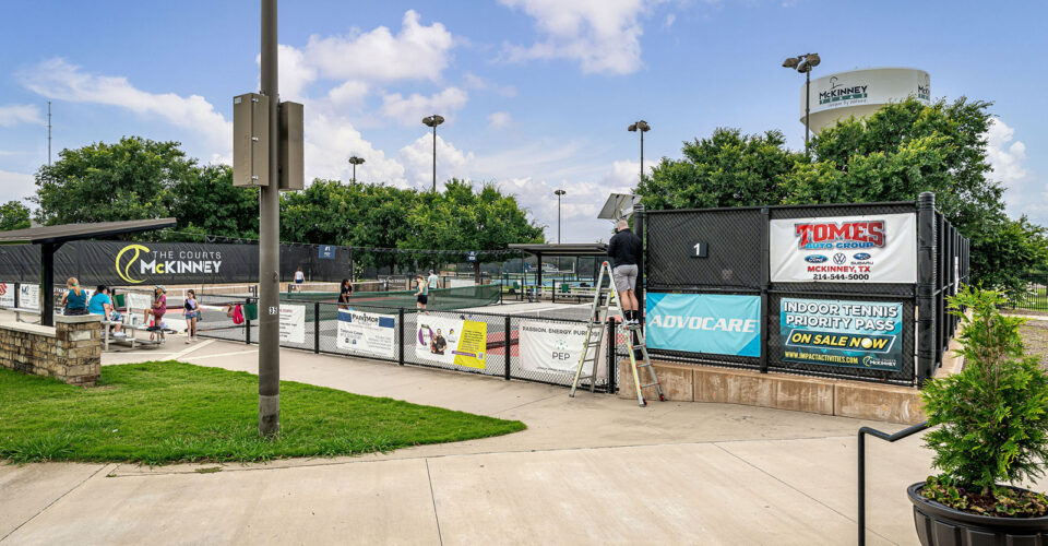 People play tennis on outdoor courts at a public recreation center, while a worker stands on a ladder near the entrance, surrounded by various banners and signs.