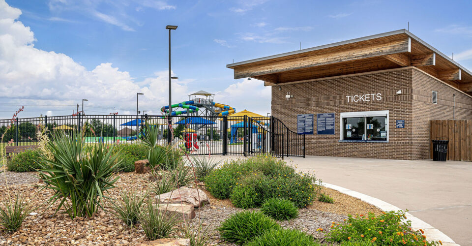 Ticket building at entrance to an outdoor water park with slides, pool, and landscaped plants in front under a partly cloudy sky.