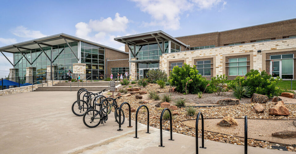 Modern school building with large windows, a landscaped entrance, concrete walkway, and bike racks with several bicycles parked.