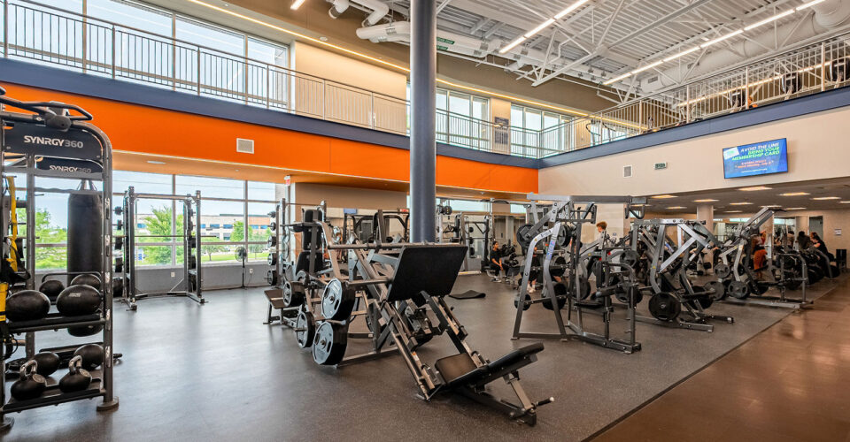 Spacious gym with various weight machines, free weights, and large windows allowing natural light; high ceilings and a mezzanine level visible.