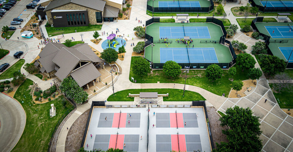 Aerial view of a sports facility with tennis courts, pickleball courts, buildings, pathways, parked cars, and landscaped green areas.