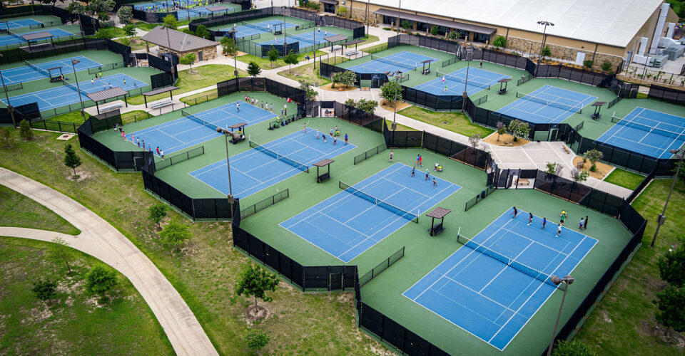 Aerial view of multiple blue tennis courts with players, surrounded by fences, pathways, and nearby buildings in a park-like setting.