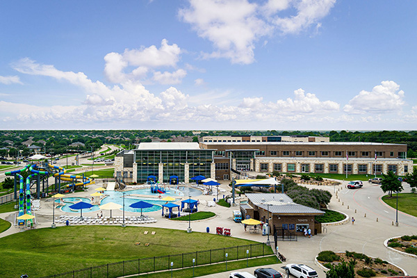 A large community center with an adjacent outdoor pool area, water slides, shaded seating, and a parking lot, set against a suburban backdrop under a partly cloudy sky.