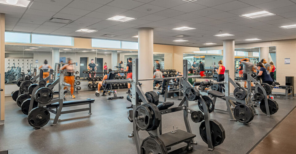 People use weightlifting equipment and free weights in a gym with racks, benches, mirrors, and bright overhead lighting.