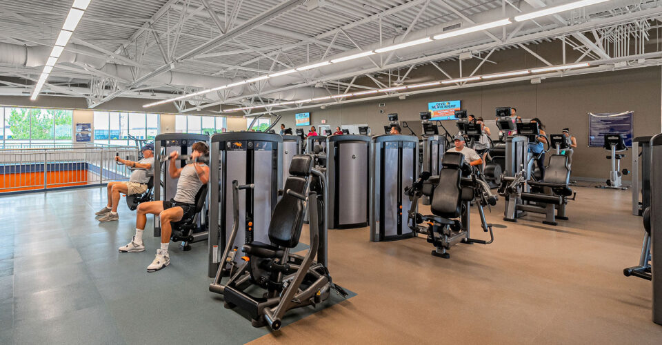 People are using weight machines and cardio equipment in a spacious, well-lit gym with high ceilings and large windows.