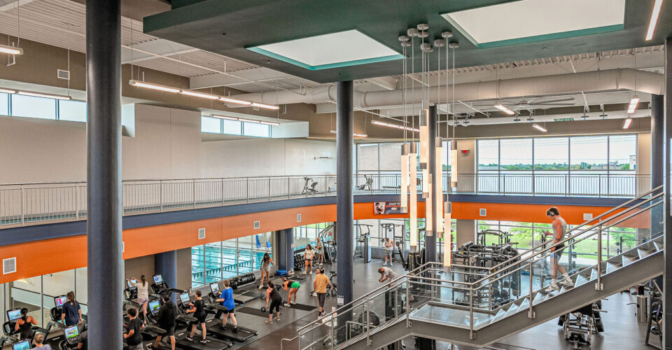 People are exercising on treadmills and bikes in a spacious, modern gym with high ceilings, large windows, and a central staircase.
