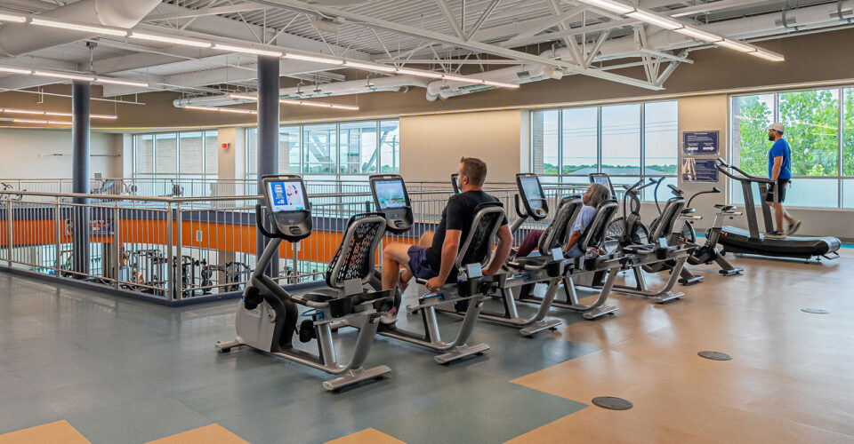 People use exercise machines in a gym with large windows and high ceilings; some are on recumbent bikes while one person uses an elliptical machine nearby.