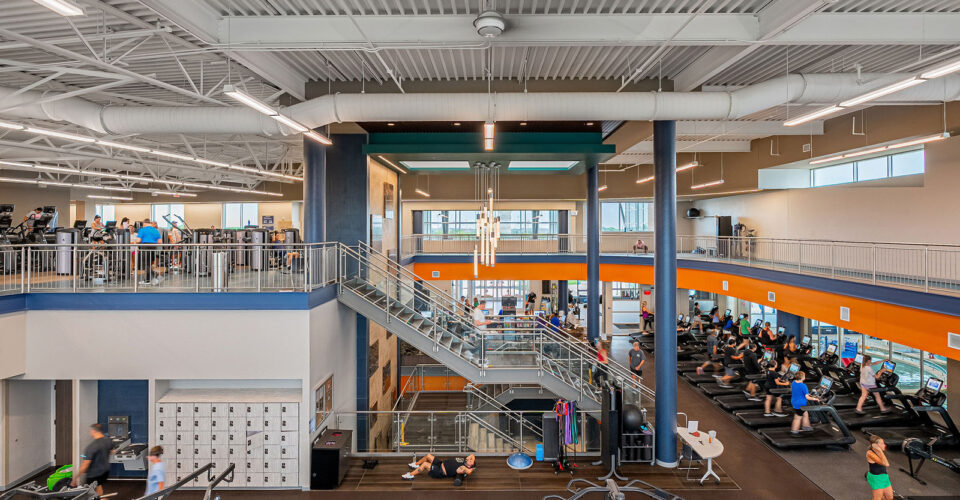 A modern gym interior with people using cardio machines, lifting weights, and walking on an upper level track. Central stairs connect two floors, and natural light fills the space.