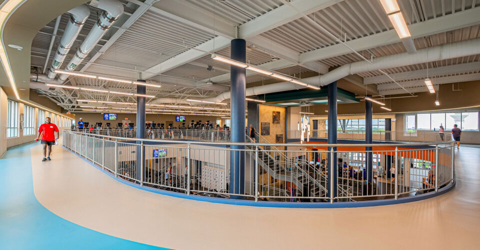 Indoor fitness center with an elevated running track, metal railings, exercise equipment, and people using the facilities. Large windows provide natural light.