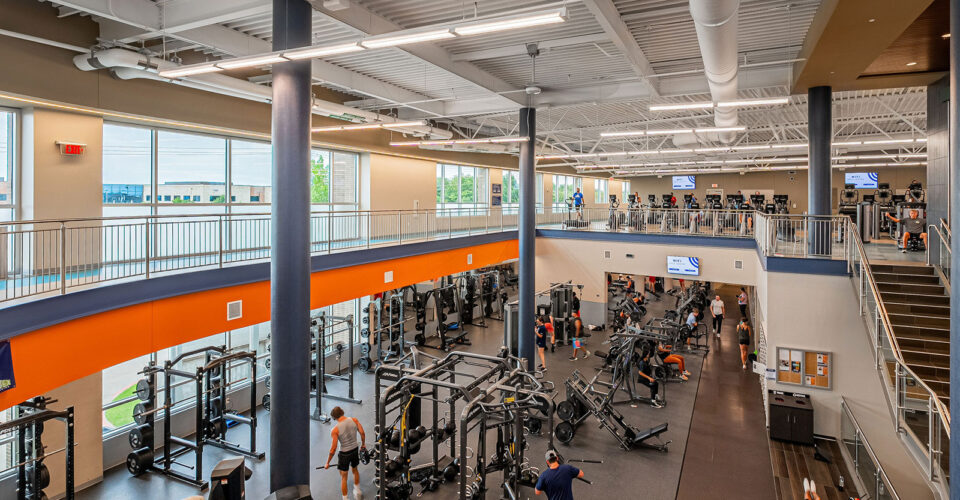 A spacious gym with strength training equipment on the lower level and cardio machines on the upper level, with several people exercising throughout the facility.