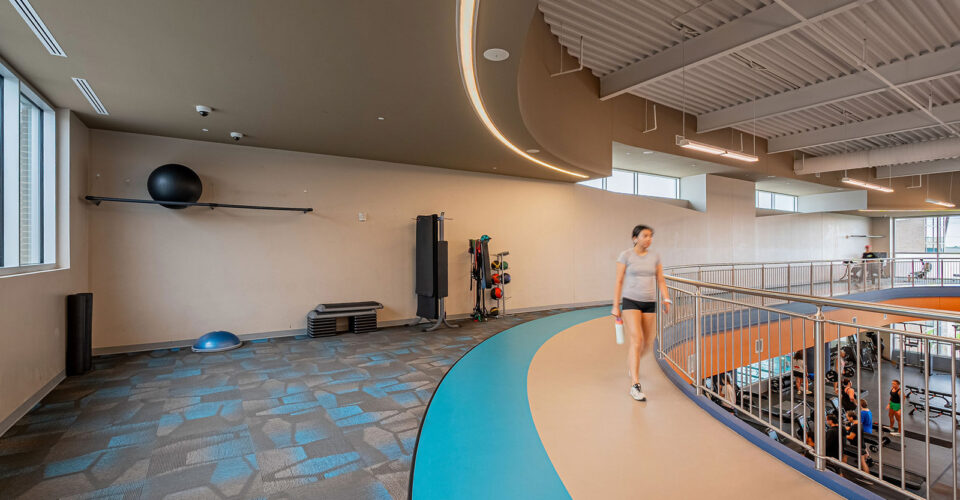 A person walks on an indoor track next to a railing, with gym equipment and windows visible in a spacious, modern fitness center.