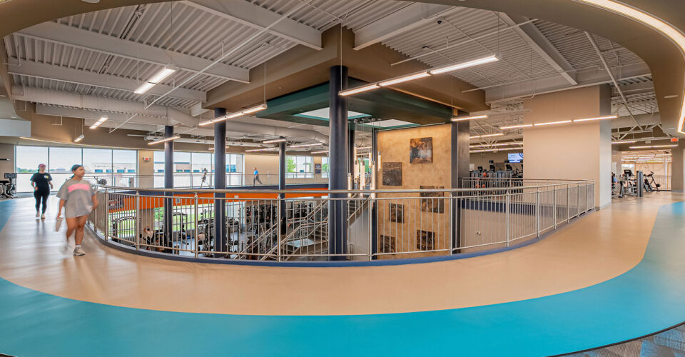 Modern indoor fitness facility with a walking track, open ceiling, stairway, gym equipment, and people exercising. Natural light enters through large windows.