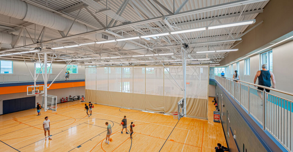 Indoor gym with a basketball court where several people are playing basketball; others are walking or running on a track above the court.