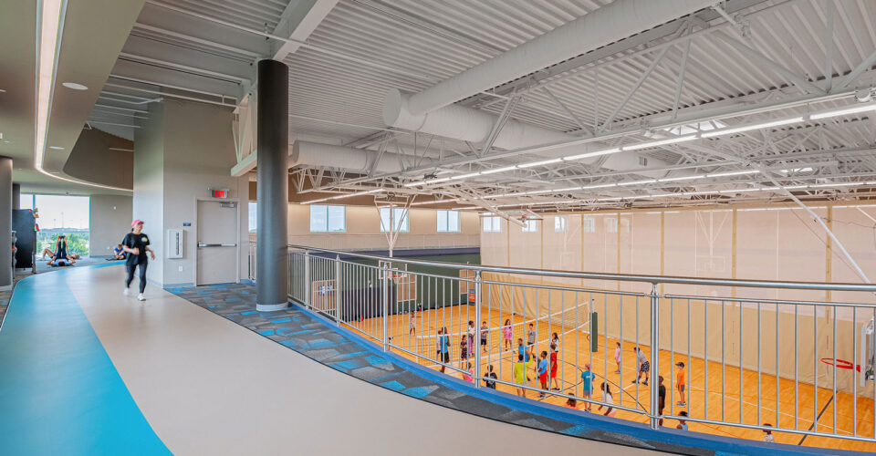 Indoor gym with people playing basketball on the court below and a person running on an elevated track above; bright lighting and modern design.