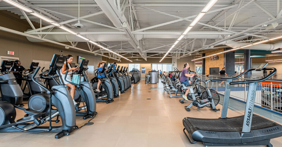 People exercise on elliptical machines and treadmills in a spacious, well-lit gym with high ceilings and large windows.