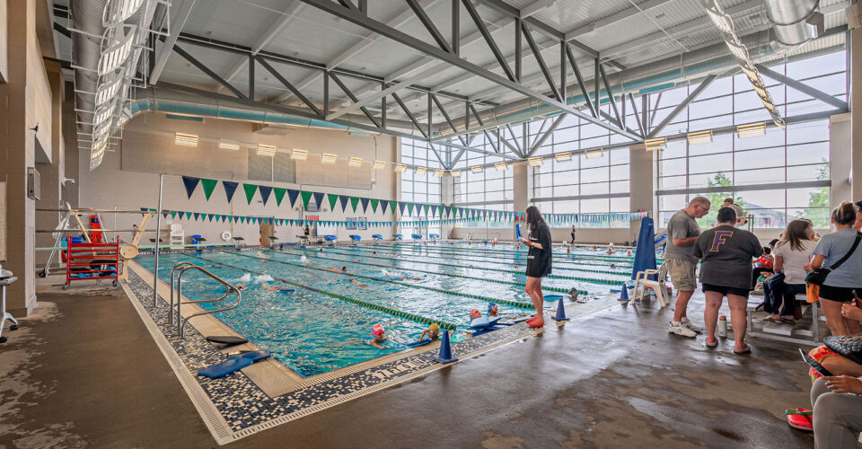 Indoor swimming pool with several people swimming, coaches and spectators on the deck, and large windows providing natural light.