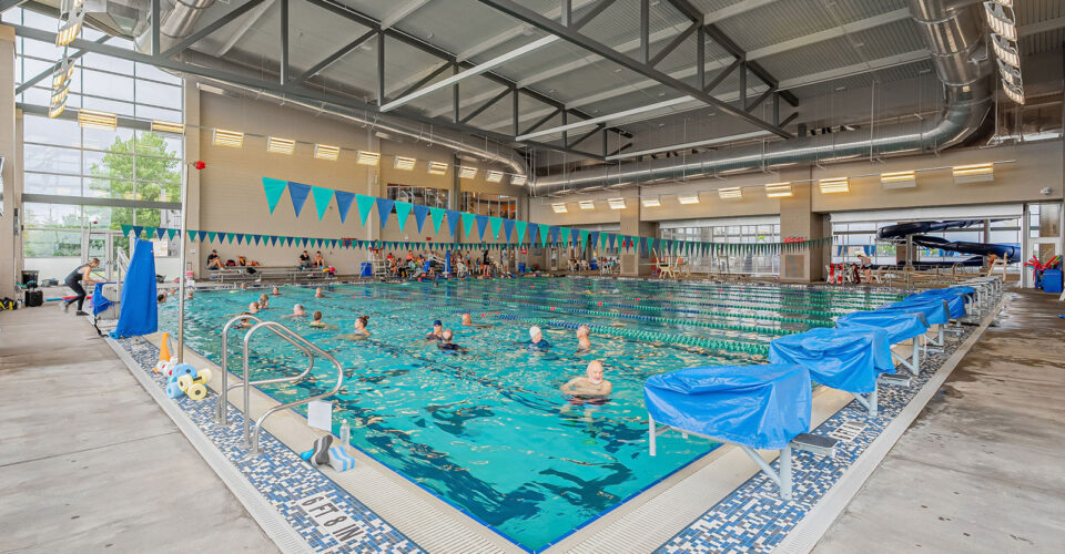 Indoor swimming pool with swimmers in multiple lanes, blue lane dividers and flags, pool equipment on the deck, and large windows providing natural light.