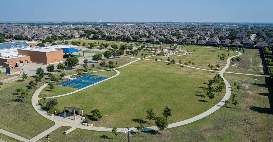 Aerial view of a large suburban park with walking paths, a basketball court, scattered trees, and adjacent residential houses.