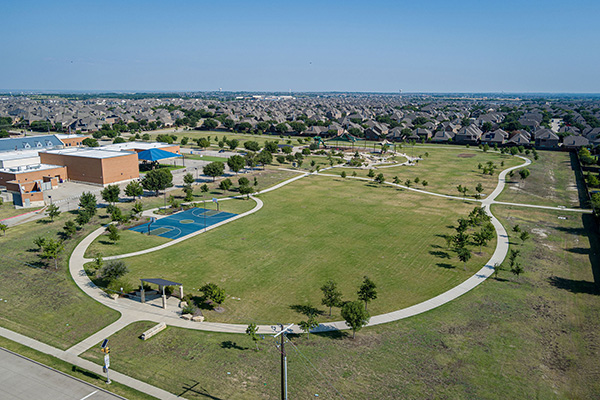 Aerial view of a suburban park with walking paths, a basketball court, scattered trees, and residential houses in the background.