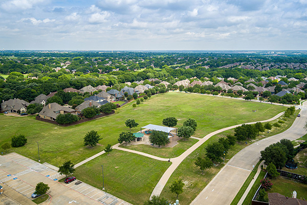 Aerial view of a suburban neighborhood with a large open grassy park, walking paths, scattered trees, a small pavilion, and surrounding houses under a partly cloudy sky.