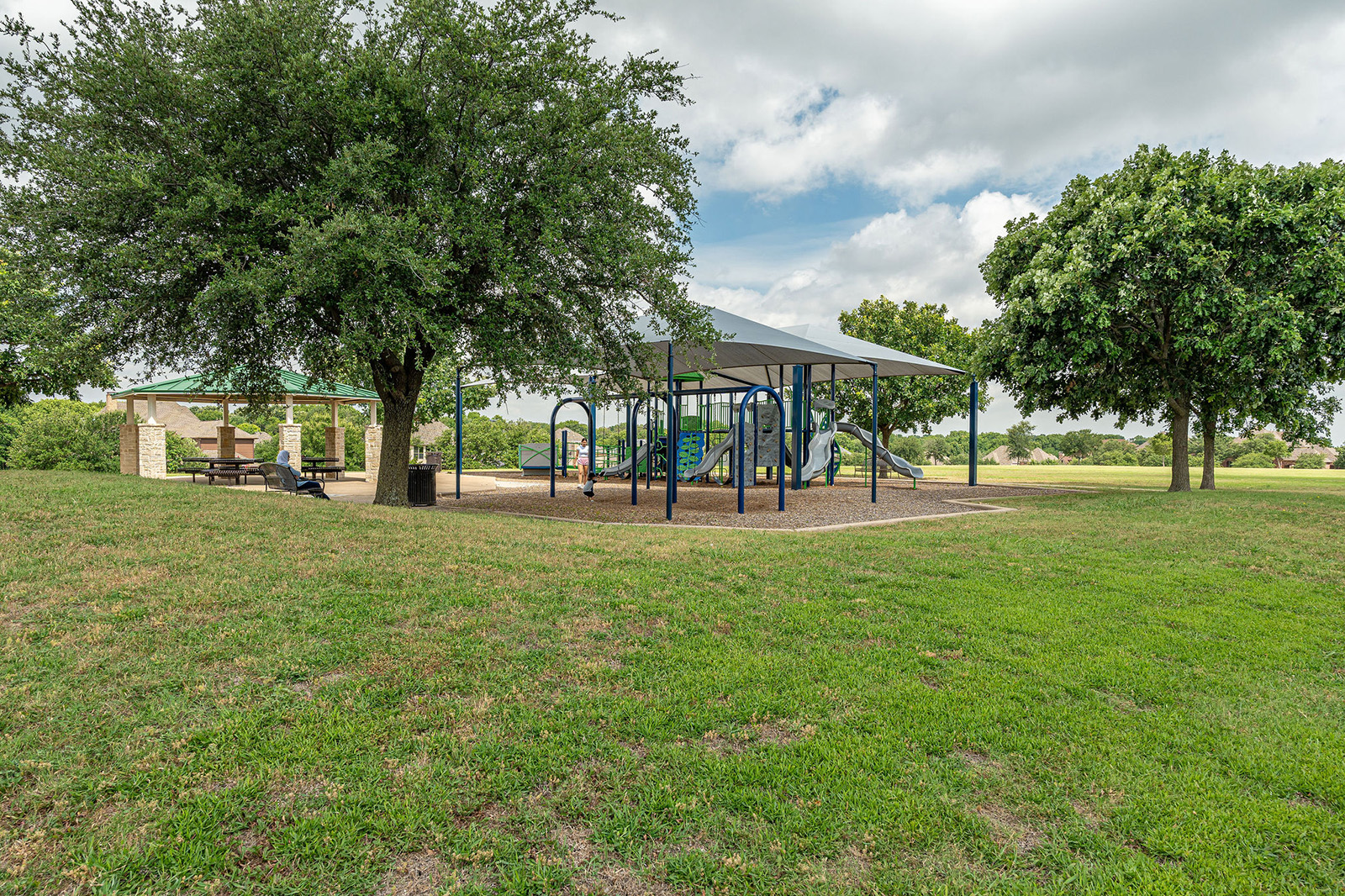 A covered playground with slides and climbing structures sits on mulch, surrounded by grass and trees, with a picnic area nearby under a pavilion.