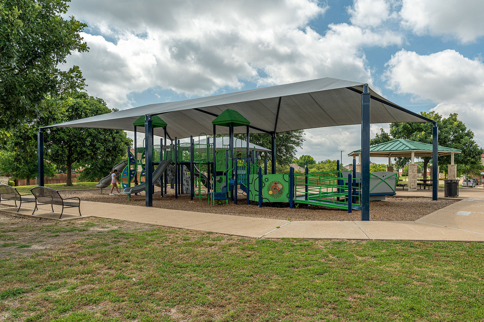 A covered playground with green play structures sits on a mulch surface, surrounded by trees, benches, and a nearby pavilion on a partly cloudy day.