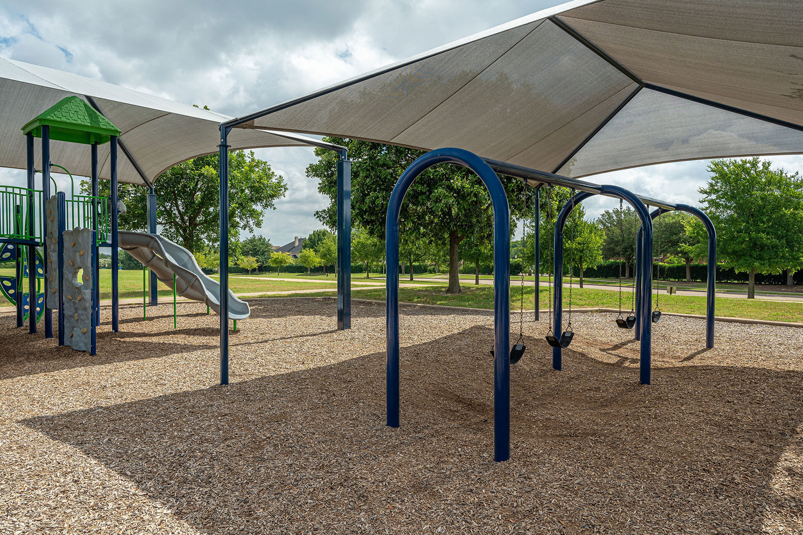 A playground with swings, a slide, and climbing equipment under large shade canopies, surrounded by trees and grass.
