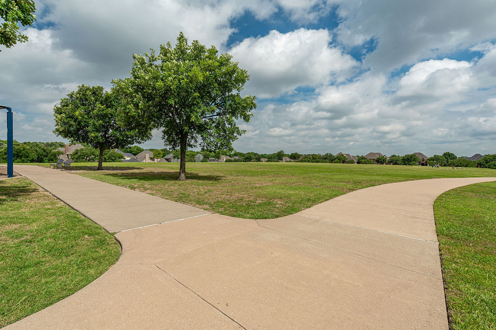 A paved sidewalk curves through an open grassy park with scattered trees and houses in the background under a partly cloudy sky.