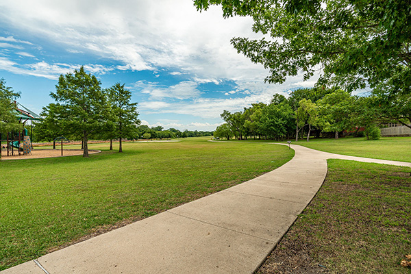 A concrete sidewalk curves through a grassy park with scattered trees under a partly cloudy sky. A playground structure is visible on the left side.