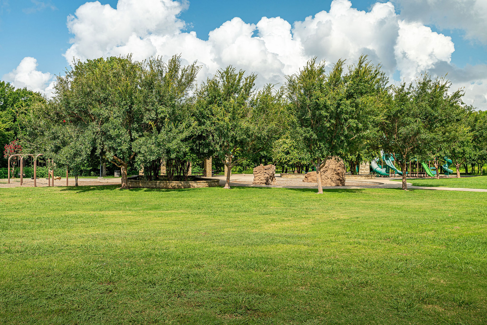 A grassy field with trees, a playground with slides and climbing structures, and a partly cloudy sky in the background.