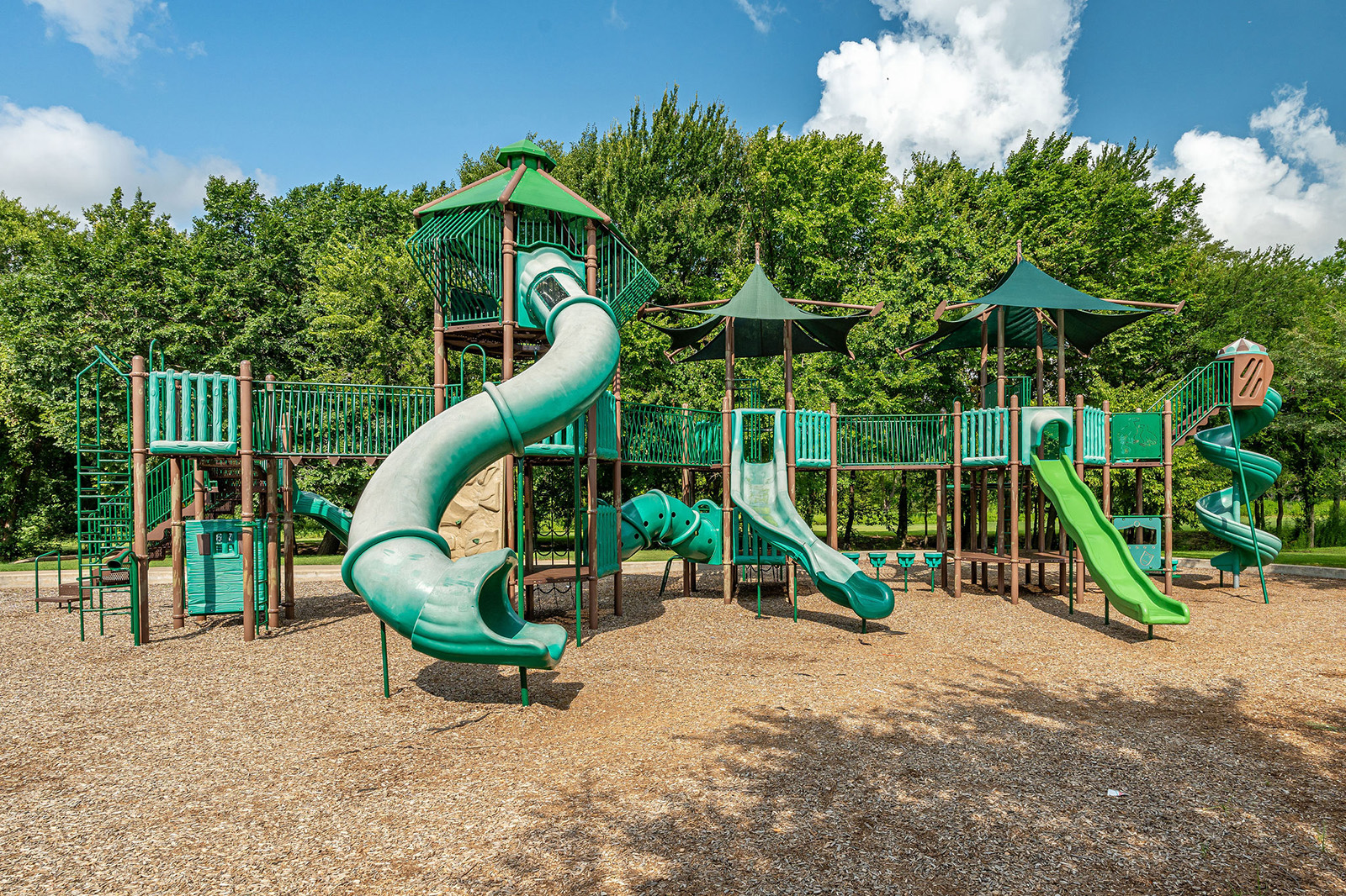 Large outdoor playground with multiple green slides, climbing structures, and towers, set on wood chip ground with trees in the background under a partly cloudy sky.