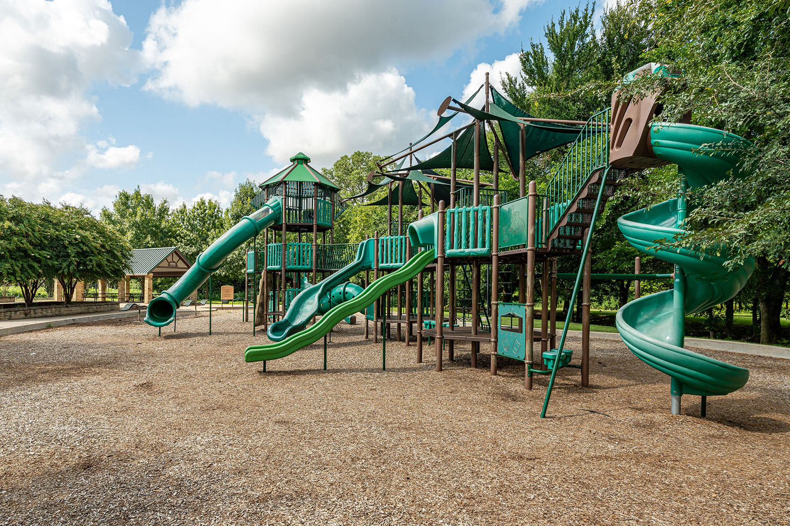A playground with multiple green slides, climbing structures, and platforms is surrounded by trees on a sunny day.