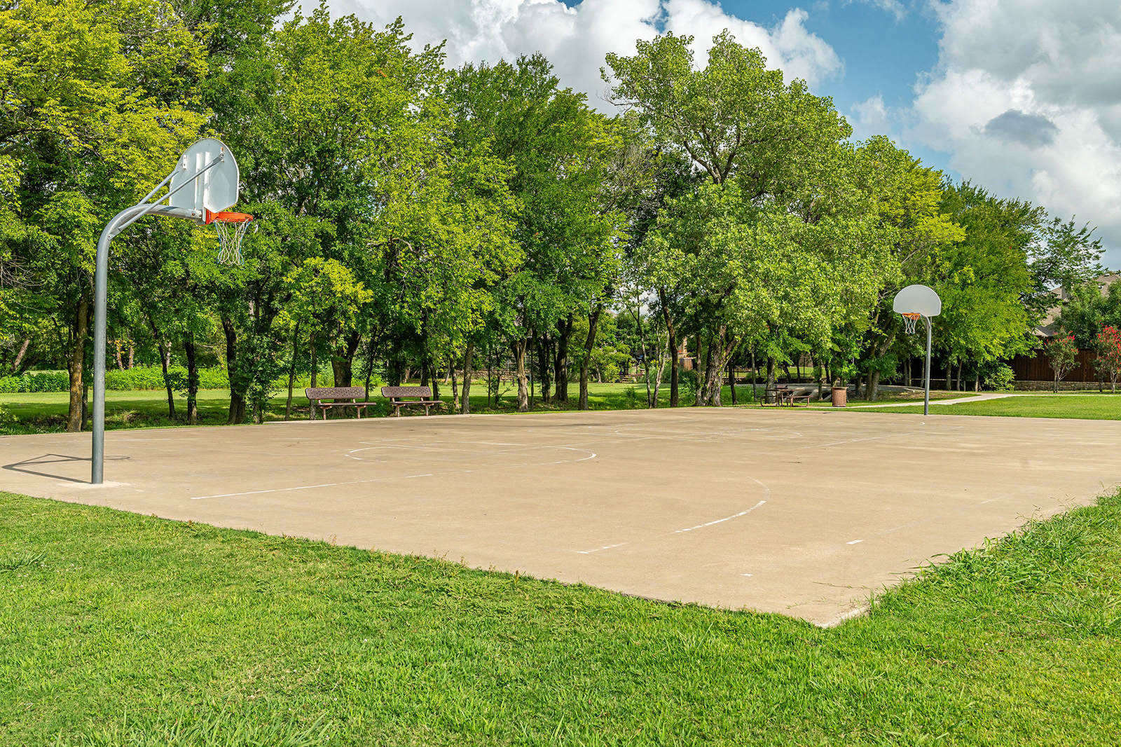 An outdoor basketball court with two hoops, surrounded by grass and trees, under a partly cloudy sky.