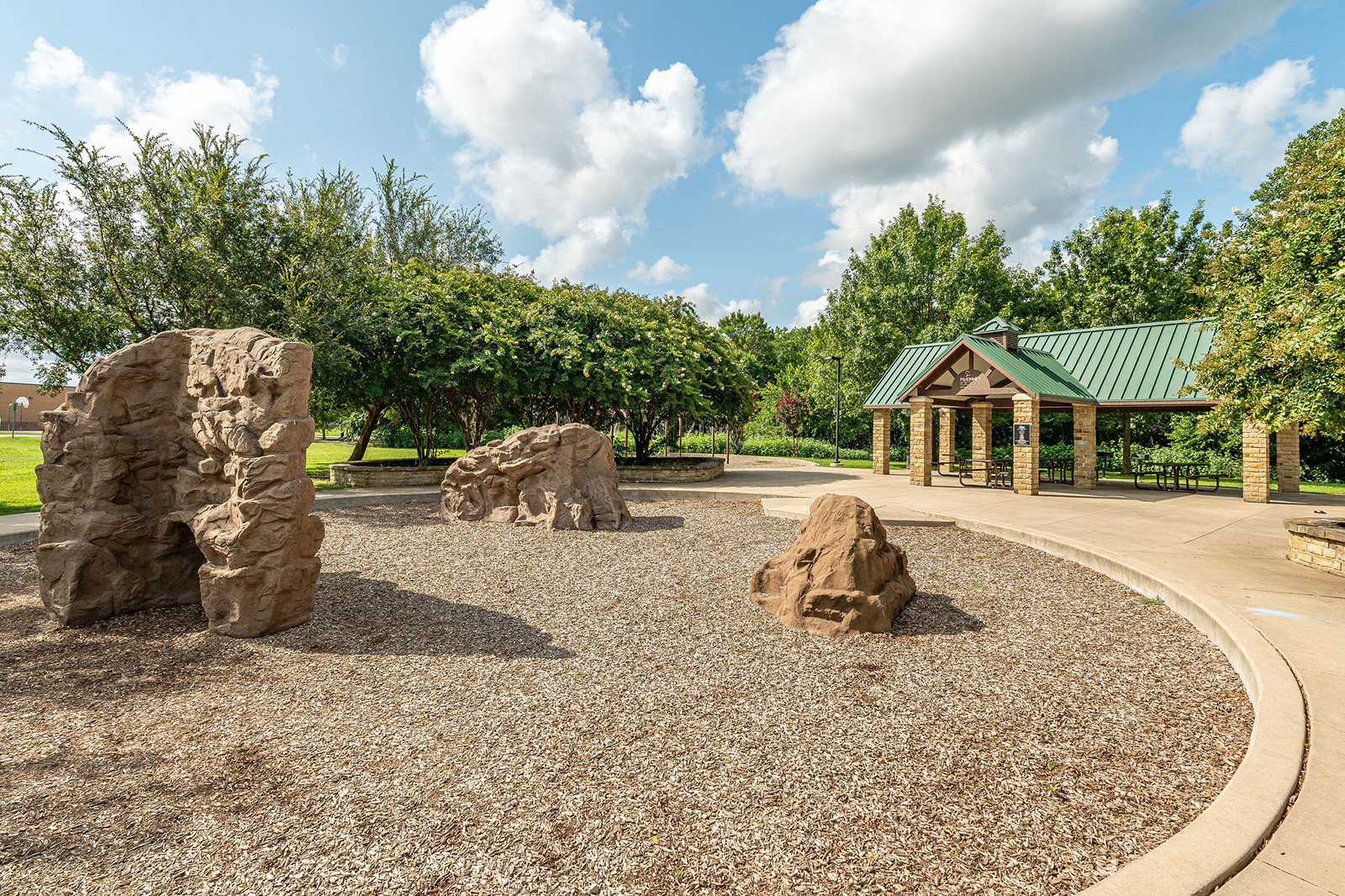 Outdoor playground area featuring artificial climbing rocks on a mulch surface, with a covered picnic pavilion and trees in the background under a partly cloudy sky.