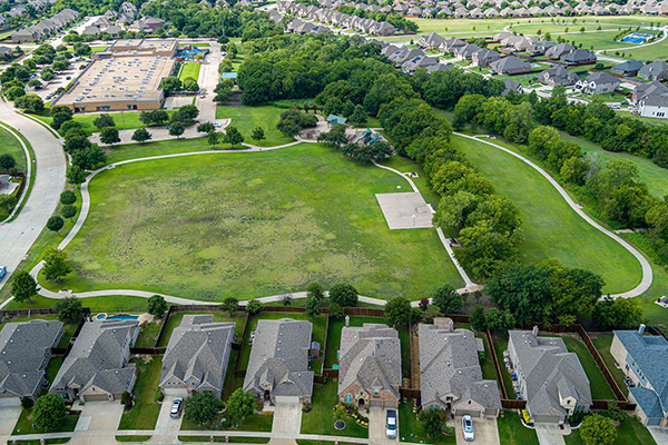 Aerial view of a suburban neighborhood with houses bordering a large, open green park area surrounded by trees and nearby parking lot.
