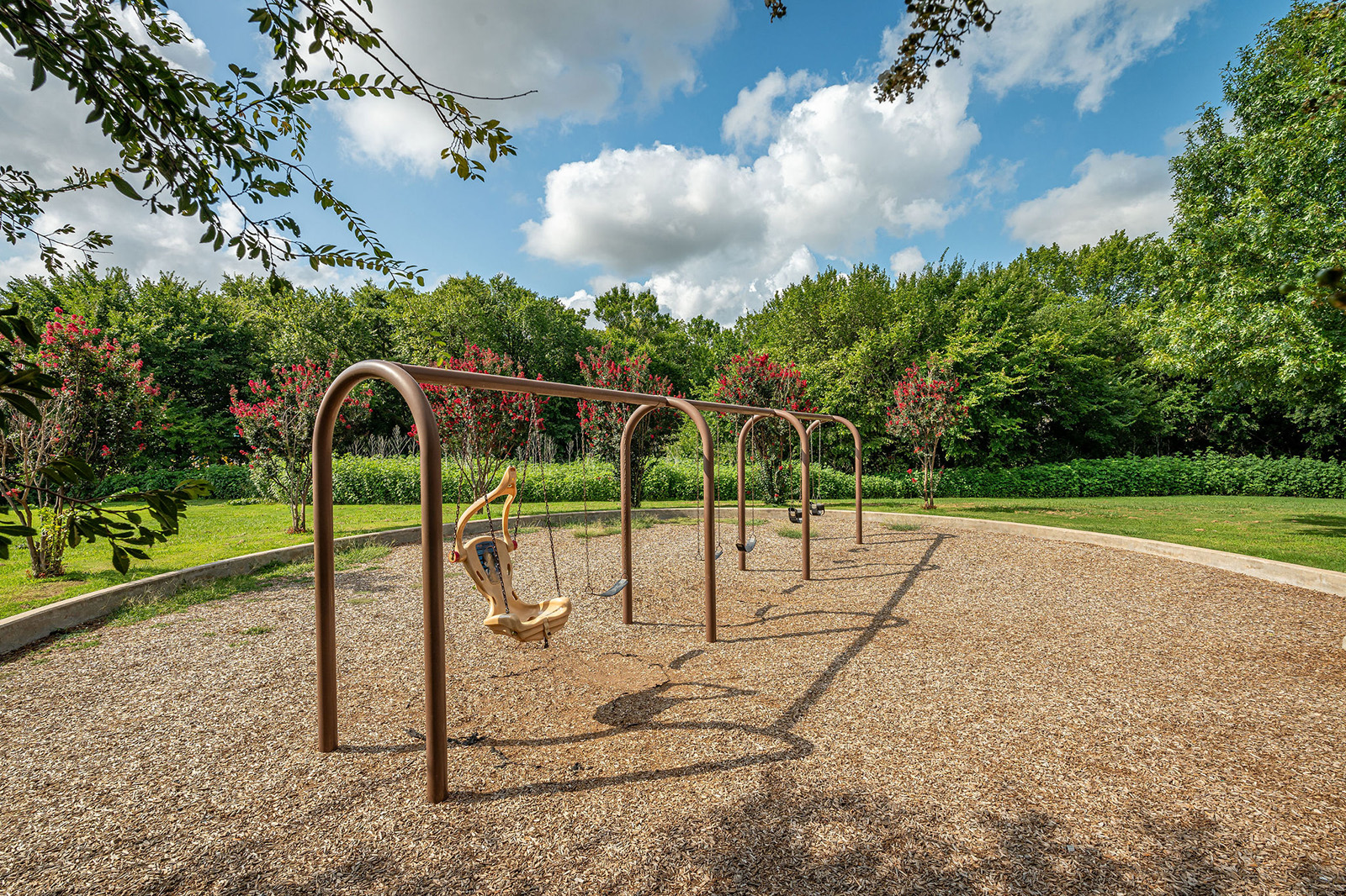 Four empty swings hang from a metal frame in a playground surrounded by grass, trees, and flowered bushes under a partly cloudy sky.