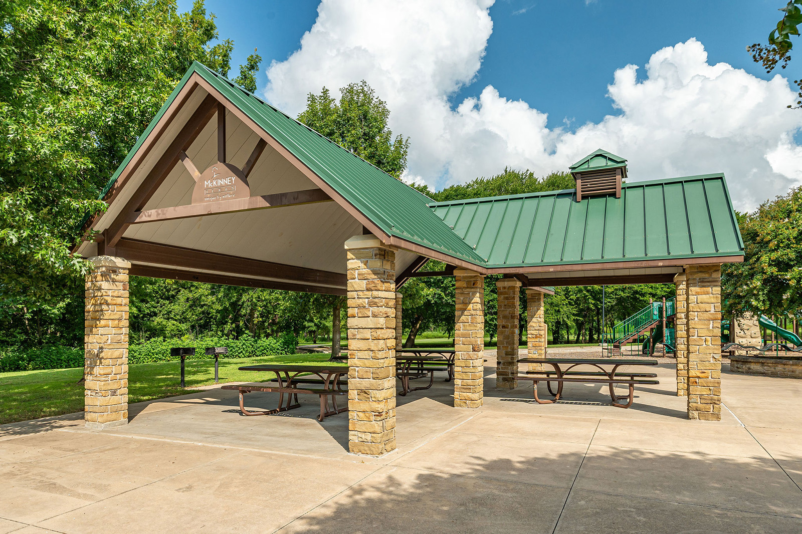 A stone pavilion with a green metal roof, picnic tables, and a grill is shown in a park setting with trees and a playground in the background.