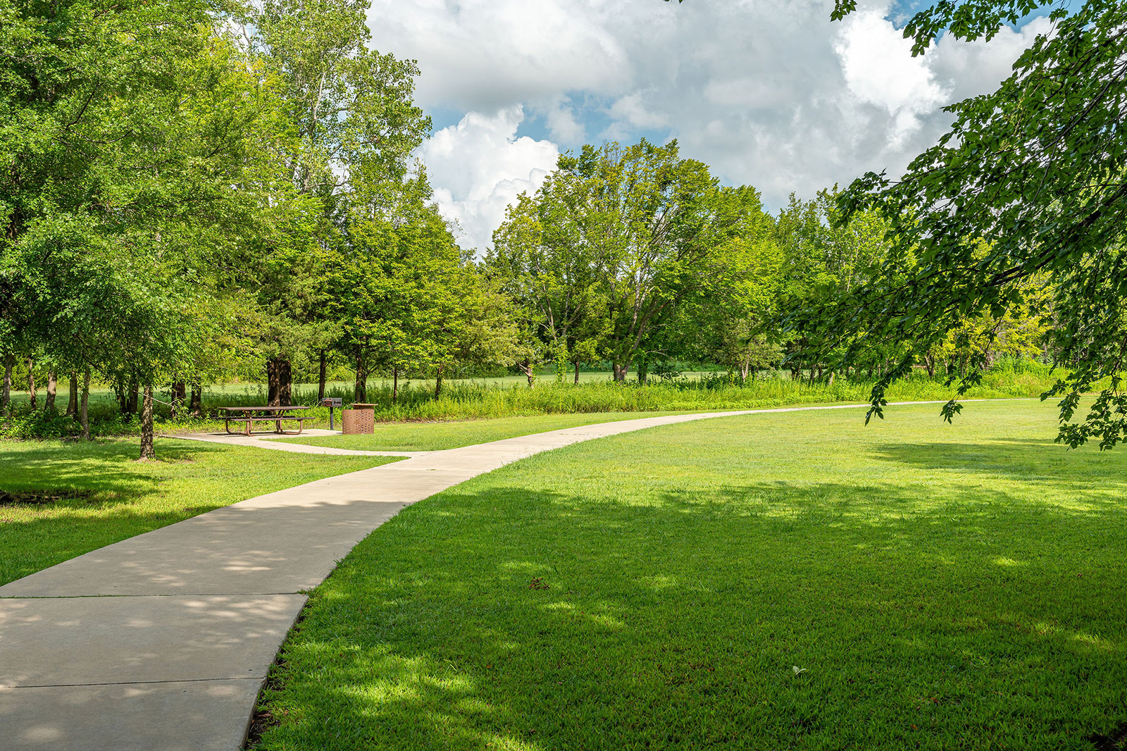 A paved walking path curves through a green park with trees, grass, a picnic table, and a trash bin under a partly cloudy sky.