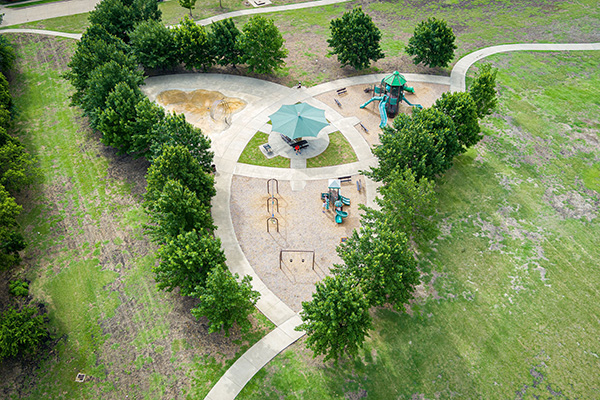 Aerial view of a small playground with climbing structures, slides, swings, a sandpit, benches, and shaded seating surrounded by trees and grass.