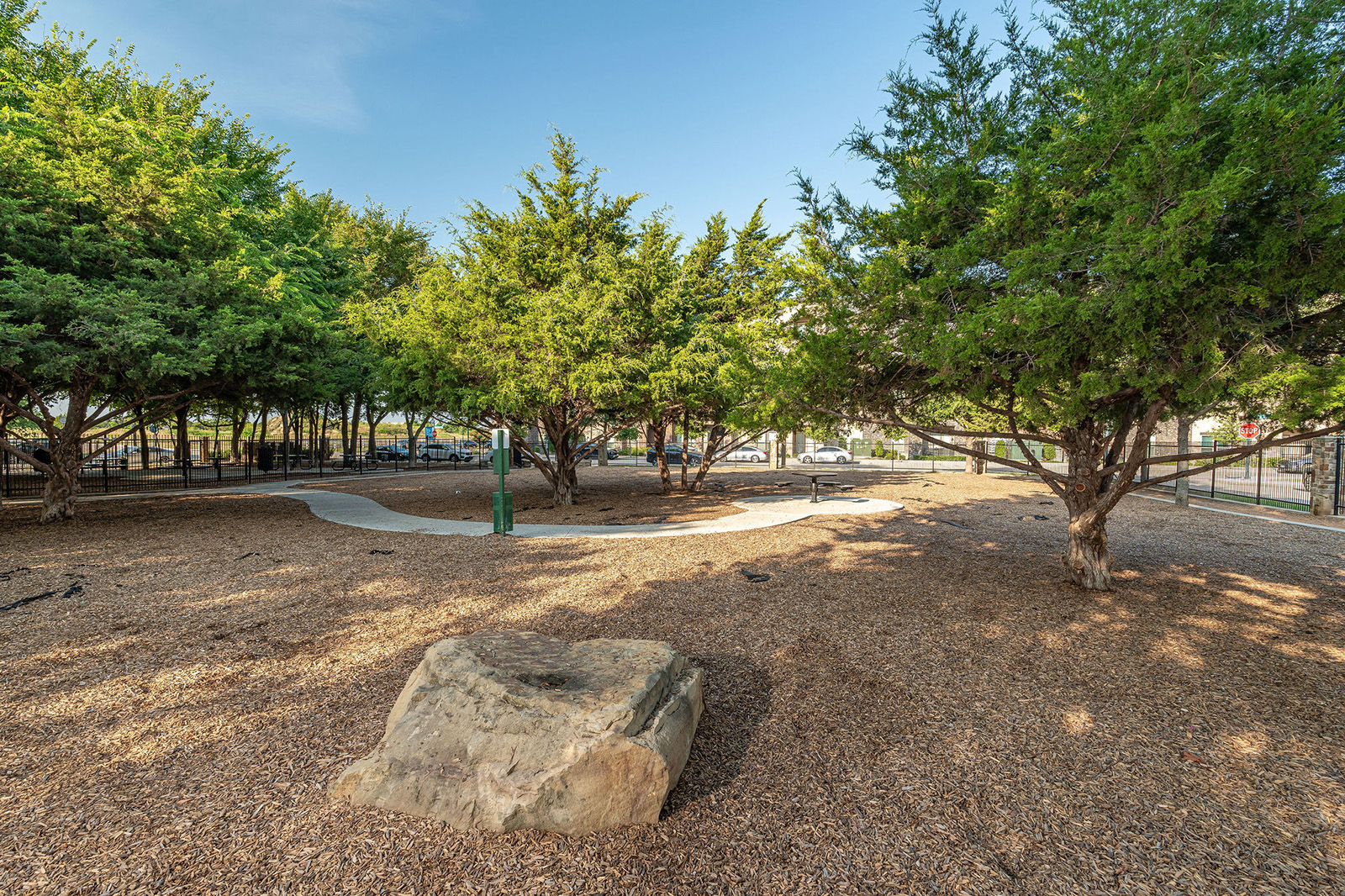 A park area with mulch ground cover, a large rock in the foreground, several trees, a winding paved path, and a water fountain in the center.