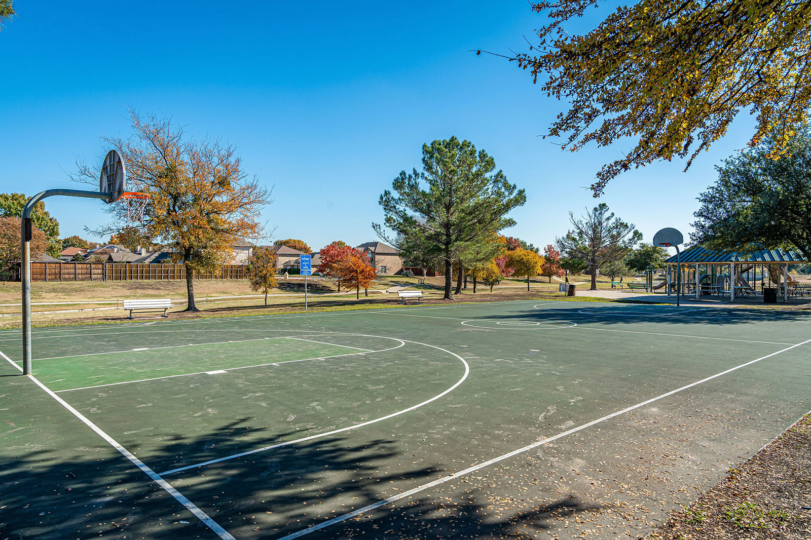 Outdoor basketball court with two hoops, surrounded by trees and benches, under a clear blue sky in a park setting.