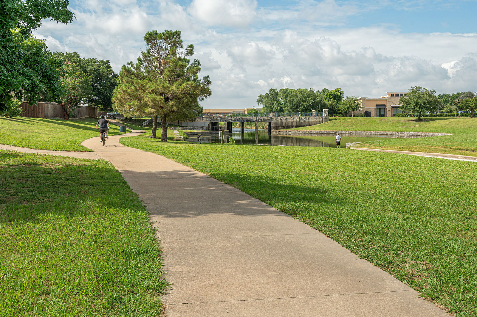 A paved walking path curves through a grassy park with a few people walking and fishing near a small bridge and pond under a partly cloudy sky.