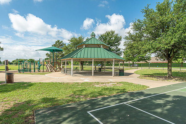 A green-roofed pavilion stands near a playground and a basketball court in an outdoor park on a sunny day.